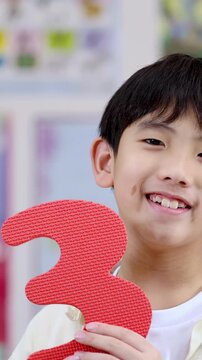 Smiling Asian Schoolboy Holding a Large Red Number Three in Classroom