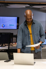 African American male reviewing papers at office desk with laptop phone charts wearing mustard knit © wavebreak3