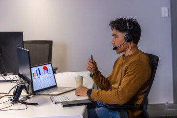 Indian man in his 20s sitting at office desk analyzing laptop charts with headset, copy space © wavebreak3