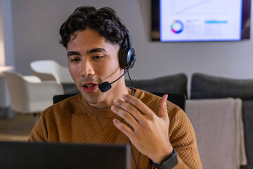Man in 20s wearing sweater, headset speaking gesturing toward laptop at home, TV chart, smartwatch © wavebreak3