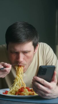 Serious young man eating spaghetti with tomato sauce for lunch while scrolling through social media on mobile phone, showing concept of phone addiction and modern lifestyle habits. Addiction concept