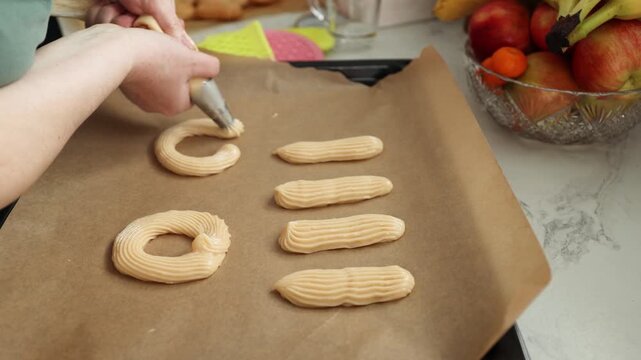 Woman piping pastry dough into ring and stick shapes on baking sheet in sunny kitchen. Baking process for cream-filled custard cakes. Home dessert preparation and filling concept. video footage video