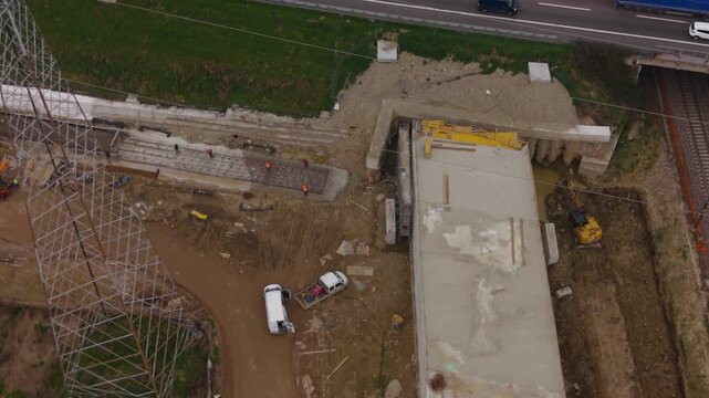 Construction Site: An aerial view of a large construction site shows ongoing work on a concrete structure, with heavy machinery and materials scattered across the muddy ground. Milan Italy