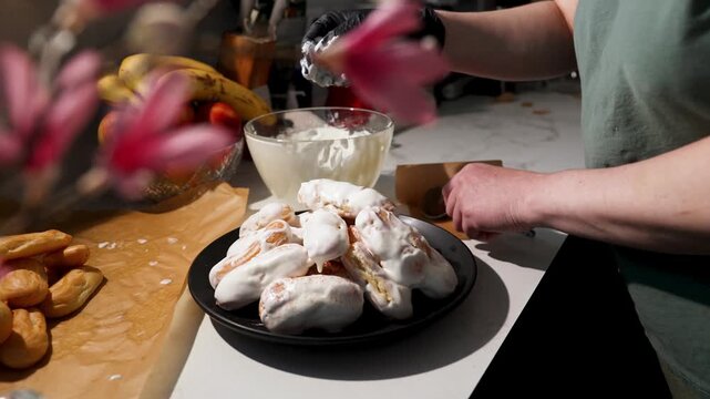 Woman pouring cream over custard donuts in sunny kitchen. Hands decorating stack of white pastries on black plate with milk drizzle and powder sprinkle. Home baking dessert preparation sequence.