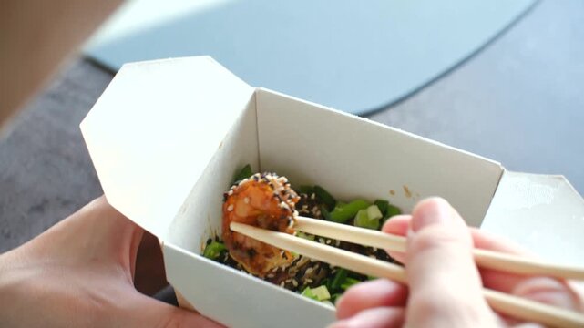 Close up woman hands using chopsticks, pick up shrimp from white takeaway box containing delicious asian meal with green onions soy sauce sesame seeds. Woman eating takeaway asian food with chopsticks