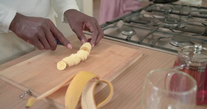 Hands are placing peeled banana on cutting board then are slicing coin rounds for cooking