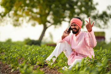 young indian farmer sitting at green agricultural field talking on smartphone