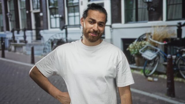 Man with hand on hip and slight smile wearing white t shirt on a cobblestone street near bicycles and row houses in city; confidence easygoing.