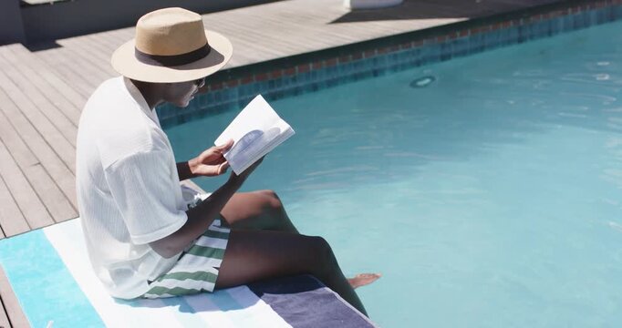 Reader is holding book on striped towel by pool, is dipping feet after pausing to cool