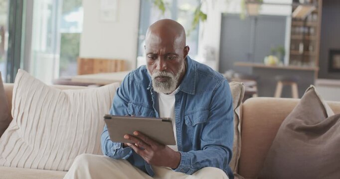 Senior African American man tapping and swiping tablet on sofa at home, referencing card for login