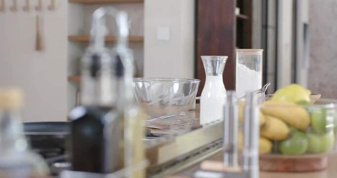Camera is panning right and pulling focus revealing mixing bowl, milk jar, oil bottles, fruit bowl