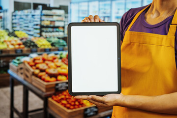 Grocery store worker in apron holding tablet with blank white screen, fresh produce aisle blurred in background, ideal for app mockup and retail promotion. © NanSan