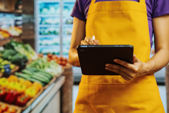Grocery store worker in yellow apron using a tablet for inventory in the produce aisle, with fresh vegetables blurred in the background.