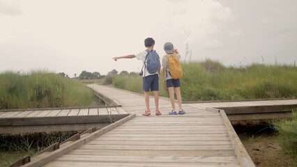 Two boys exploring outdoors, standing on a boardwalk at a crossroads, choosing between diverging paths, wearing backpacks and pointing ahead, discovering nature's direction © DawDunia