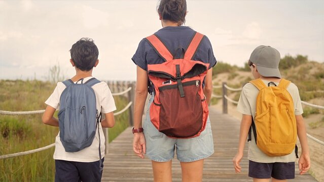 Family walking on wooden boardwalk to the beach, mother and two sons with backpacks enjoying summer nature travel adventure and vacation discovery