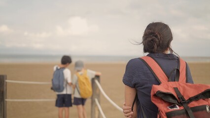 Woman with backpack stands on a wooden fence path watching two children at the beach edge, facing the vast ocean and sky—summer family escape, carefree seaside moment © DawDunia