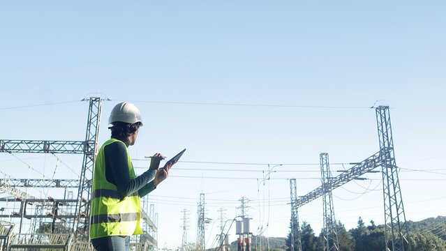 Electrical engineer checking power grid infrastructure, inspecting high voltage electricity substation equipment with a digital tablet for maintenance and energy distribution management