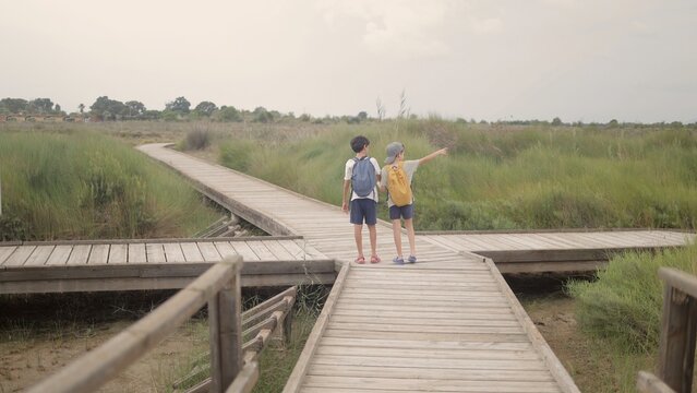 Two young boys wearing backpacks standing at a wooden boardwalk intersection, one pointing encouragingly, discovering new paths in a sprawling natural swamp landscape
