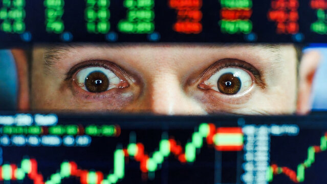 A person stares intensely at a stock market screen displaying fluctuating red and green graphs. They appear anxious.
