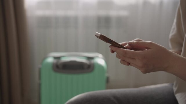 Close up of female hands using a smartphone to book a taxi online, preparing for departure with a suitcase in the background, symbolizing travel and modern transportation booking.