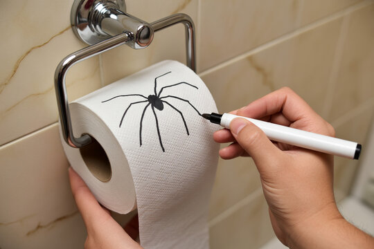 A person's hands draw a large black spider onto the white paper of a toilet roll while it is mounted on a chrome holder in a bathroom.