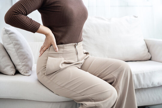 Asian woman suffering from lower back pain while sitting on a sofa. Young female touching her aching back, feeling muscle strain, spinal problem, or office syndrome symptoms in the living room