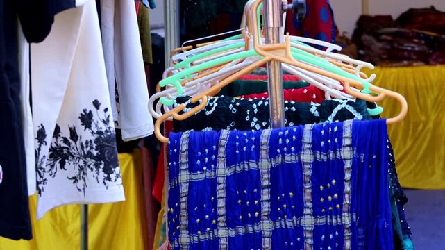 Colorful garments and scarves displayed on plastic hangers at a market stall, featuring patterned fabric and embroidered textiles hanging on a rotating rack.