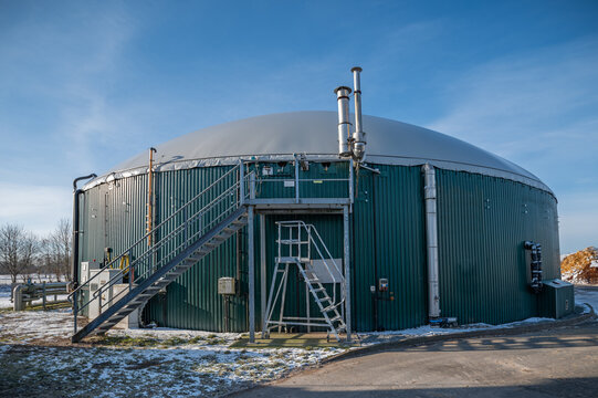 Exterior view of agricultural biogas plant with large anaerobic digester tank and domed membrane roof equipped with external piping, ventilation stacks and access platform with stairs for maintenance.