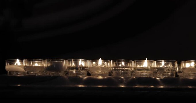 Burning tealights in clear holders on a marble altar ledge, steady view shows gentle flicker and warm glow as melted wax and tiny wicks catch the only light inside the church gloom