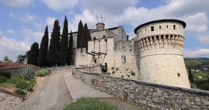 Brescia castle approach with cypress and bastion in view, camera pans right to left from the lifting bridge to Torre Mirabella as tricolor and eu flags wave under a clouded blue sky