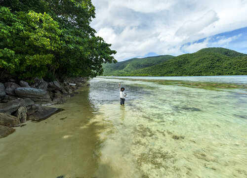 Aerial view of a person standing in the shallow, turquoise waters of Bobby Beach, framed by lush greenery and distant hills, Karimunjawa, Central Java, Indonesia.