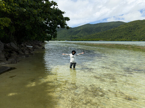Aerial view of a person standing in the shallow, clear waters of Bobby Beach with arms outstretched, framed by lush greenery and distant hills, Karimunjawa, Central Java, Indonesia.