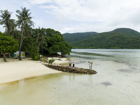 Aerial view of the tranquil Bobby Beach, where white sand meets clear blue water, contrasting with lush green vegetation and two girls playing together, Karimunjawa, Central Java, Indonesia.