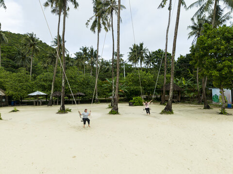 Aerial view of light sand and tall palm trees framing people on swings at Bobby Beach, Karimunjawa, Central Java, Indonesia.