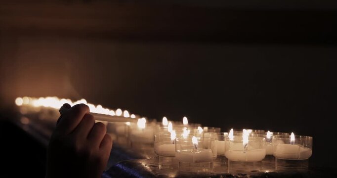 Prayer candles and hands lighting tapers on marble altar, in a dim church a row of candles flickers while hands touch long candles to the small flames and wax pools in clear cups as thin smoke
