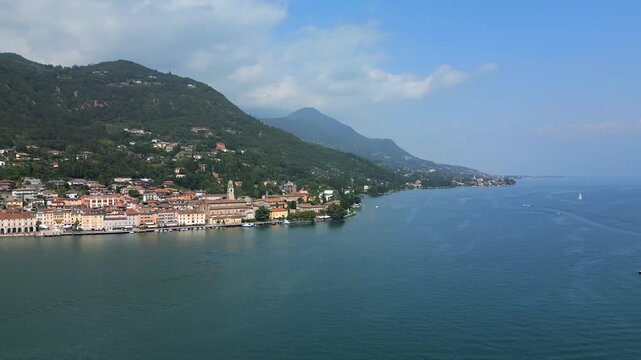 Hovering drone yaw panorama over Salo on Lake Garda, camera pans right to left above marina and promenade with pastel buildings and duomo di salo beneath green alpine foothills as bright summer sun