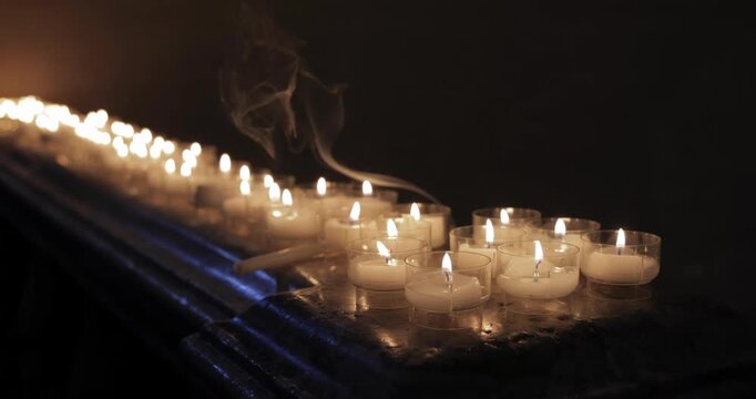 Votive candles on a marble altar in a dim church interior, static camera holds on flickering flames in clear cups as warm light reflects on stone and thin smoke drifts upward