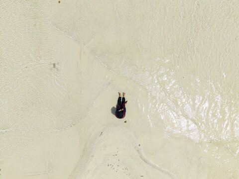 Aerial view of a person sitting on the shore, surrounded by the light, soft sand meeting the clear water at Cemara Kecil Island, Karimunjawa, Central Java, Indonesia.