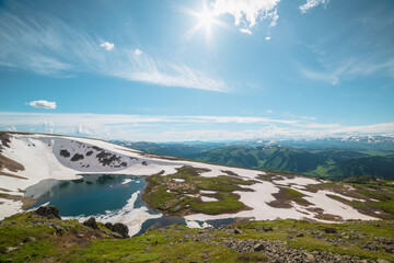 Scenic sunlit landscape with alpine lake in rocky snowy cirque near stone hill top in sunny day during thaw. Ice floats in mountain lake among rocks with view to forest mountain range under bright sun © Daniil