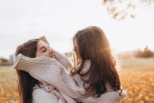 Mother and daughter wearing cozy knitted sweaters share warm embrace outdoors during golden-hour in autumn. Glowing intimate family portrait. Maternal bond, love, mother&rsquo;s day. Copy space.