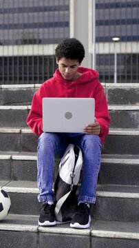 Vertical video: Opening laptop, man in red hoodie on plaza steps typing work, backpack, soccer ball