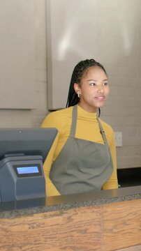 Vertical video: Greeting cashier wearing gray apron, on customer arrival at cafe counter, using POS