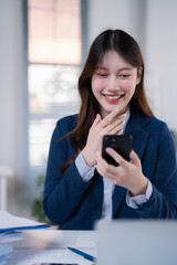 Obraz premium Young Asian businesswoman smiling brightly, looking at her smartphone while working in a modern office, expressing happiness and success in her professional communication