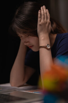 Young Asian woman at desk clutching her head and covering her ears, overwhelmed by work stress and headache, exhausted and frustrated by pressure and burnout in dim indoor light