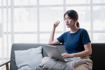 Obraz premium Young Asian woman sitting comfortably on a sofa, looking pensively at the camera while working or studying using her laptop in a bright modern living room