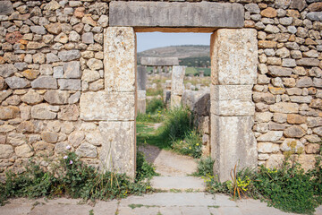 Stone archway entrance framed by weathered stone walls leading to a grassy pathway in an ancient archaeological site surrounded by hills and vegetation