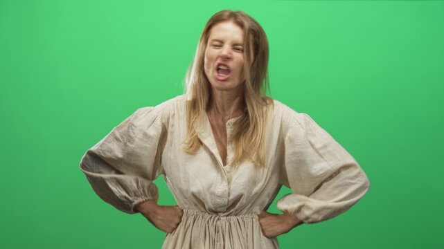 Young blonde woman with hands on hips and mouth open speaking toward camera in a green studio, scowling and leaning forward; frustration assertiveness tension.