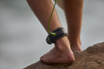 Close-up of a surfer&rsquo;s ankle secured with a safety leash while stepping toward the ocean, capturing the anticipation of entering the waves. The image highlights the connection between surfer and board