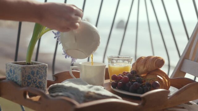 Hand pours coffee into mug on wooden tray. Tray holds fruits, croissants, and small potted plant. Morning light shines through balcony railing. Scene feels calm, relaxing, and inviting