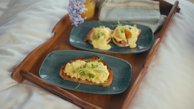Wooden tray holds two plates of creamy eggs benedict on toast. Garnished with fresh herbs and edible flowers. Soft morning light highlights rich textures and colors